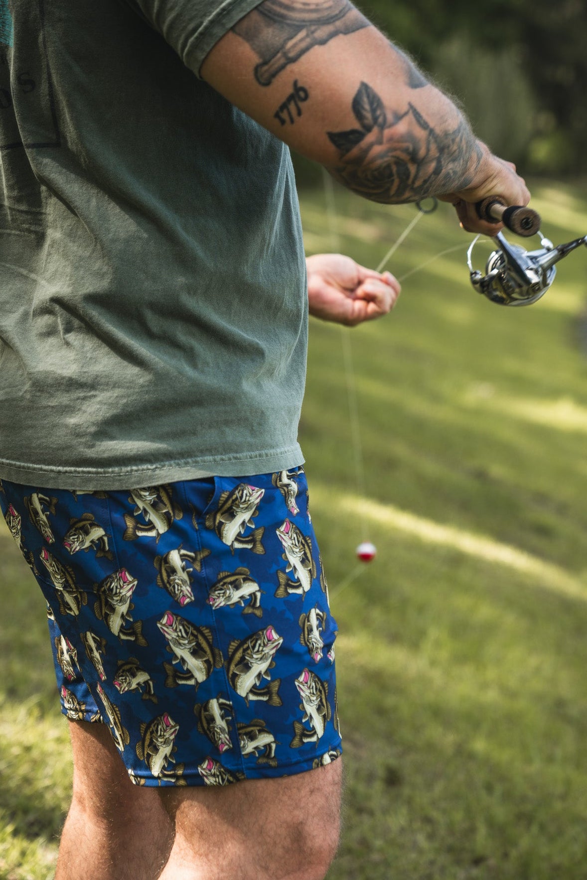 A tattooed person fishes outdoors, wearing Mucker Bucket Men's Performance Athletic Shorts | 5.5"- Big Bass Daddy. They're holding a rod with a red and white bobber, surrounded by grass and trees in the background.