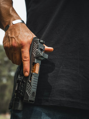 A person with a black shirt and bracelet holds a Born United Private Label handgun, their stance urgent. The accessory attachment gleams, while the greenery in the blurred background contrasts sharply, echoing the tension of an ending moment.