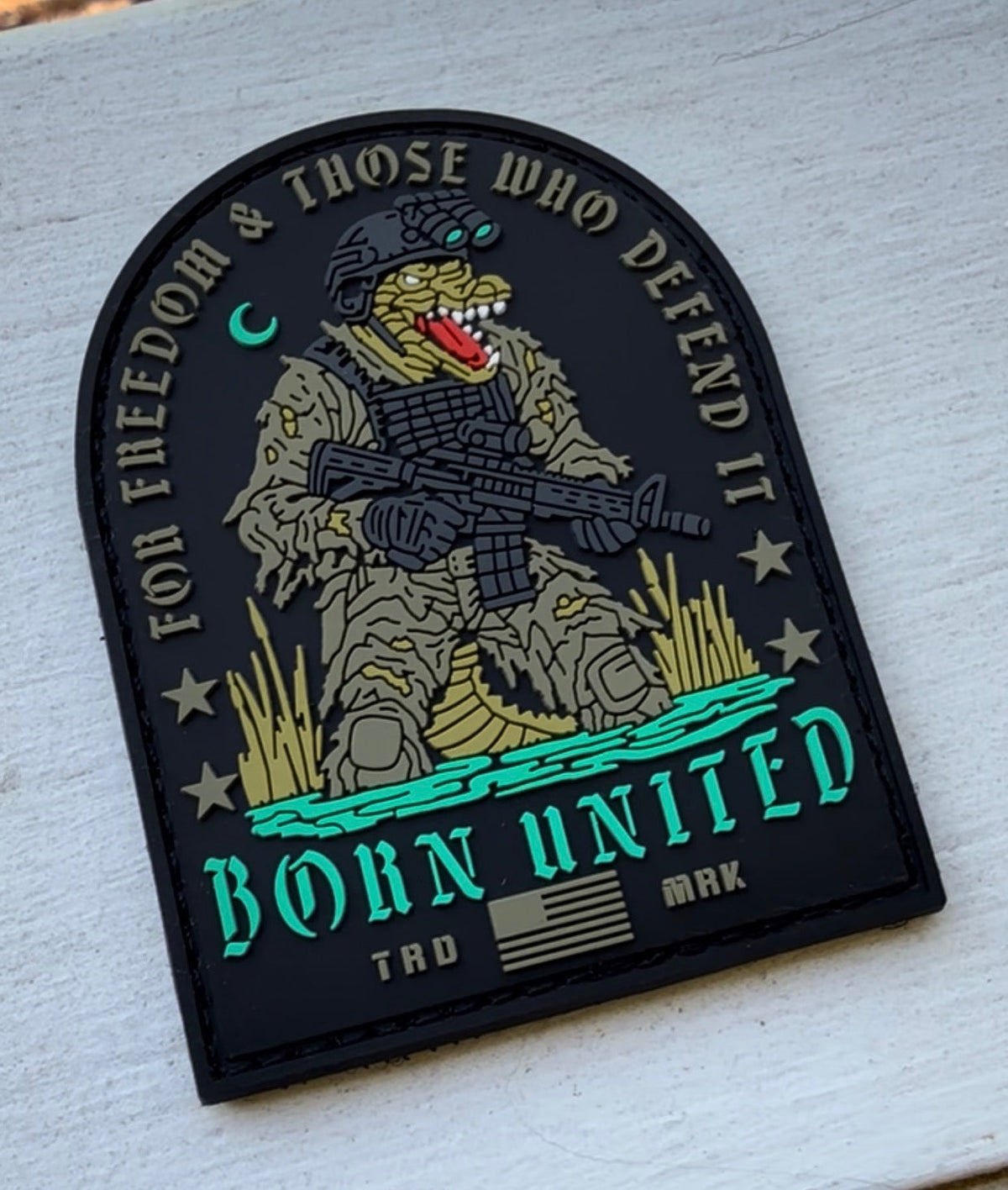 The TactiGator PVC Patch by Born United Private Label features an alligator in combat gear with rifle, the text "FOR FREEDOM & THOSE WHO DEFEND IT" and "BORN UNITED," blue water, grass, and a small American flag at the bottom.