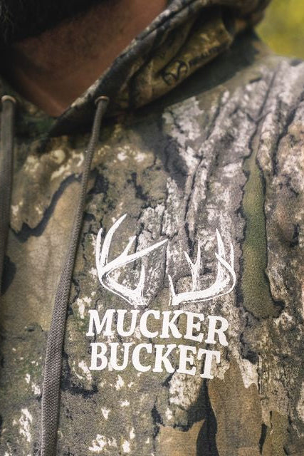 A person wearing The Rut Realtree Premium Hoodie, featuring a white logo with deer antlers and the text MUCKER BUCKET on the chest.