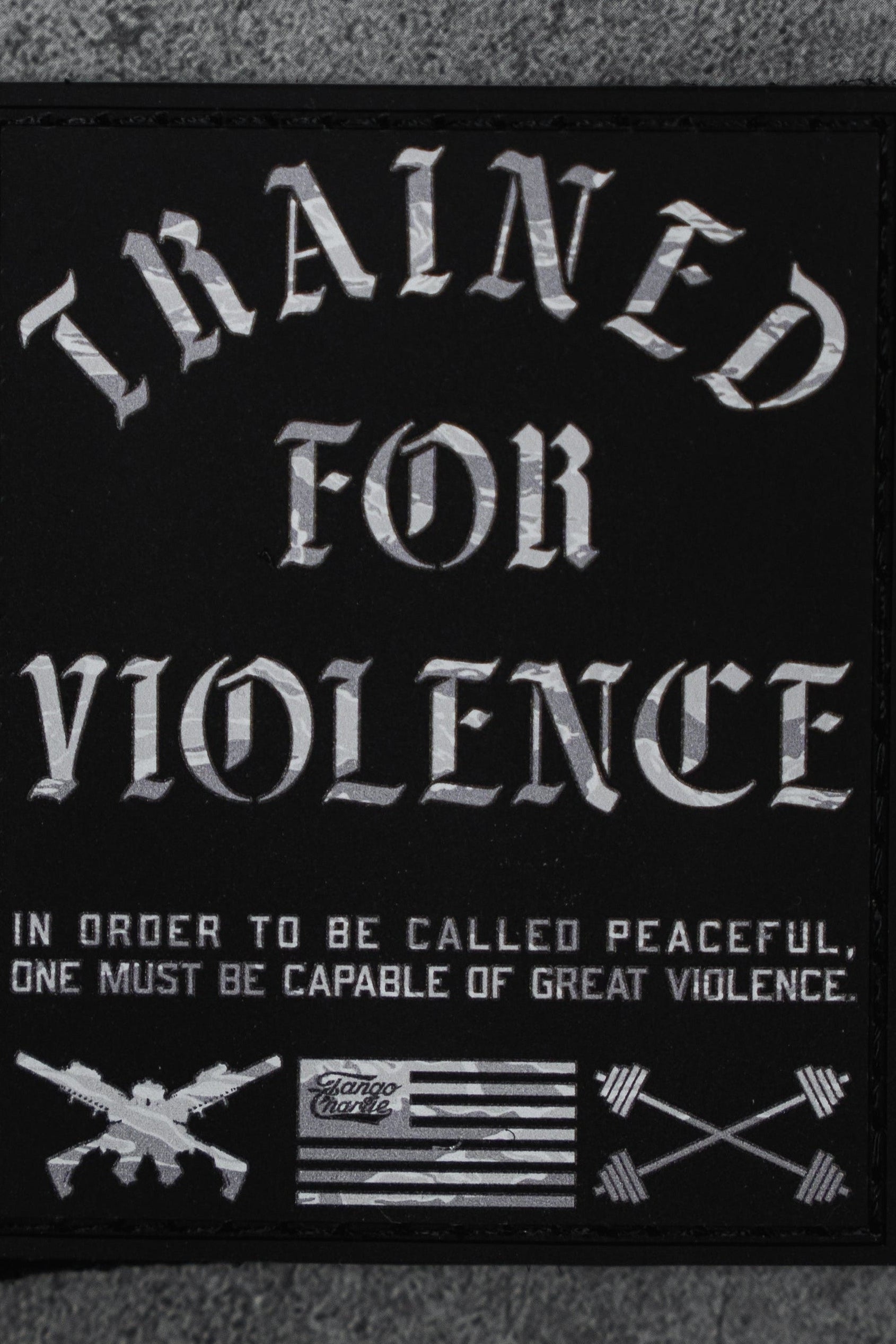 The Trained For Violence - Patch by Tango Charlie Apparel is a 3" black Velcro patch featuring bold white "TRAINED FOR VIOLENCE" text, crossed rifles, arrows, a black-and-white flag, and a striking 3D PVC design.
