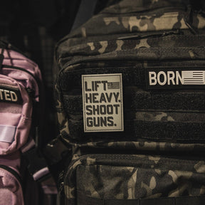 A close-up of a black camo backpack with the LHSG - Patch by Tango Charlie Apparel, featuring "LIFT HEAVY. SHOOT GUNS." in bold Velcro, alongside a 3D PVC patch reading "BORN" with an American flag. A pink bag appears on the left.