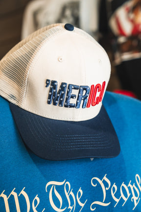 The Born United Private Label ’Merica Trucker Snapback, a white and blue trucker hat with “MERICA” across the front, is displayed atop a blue shirt with decorative script text. This breathable snapback design is ideal for everyday wear.