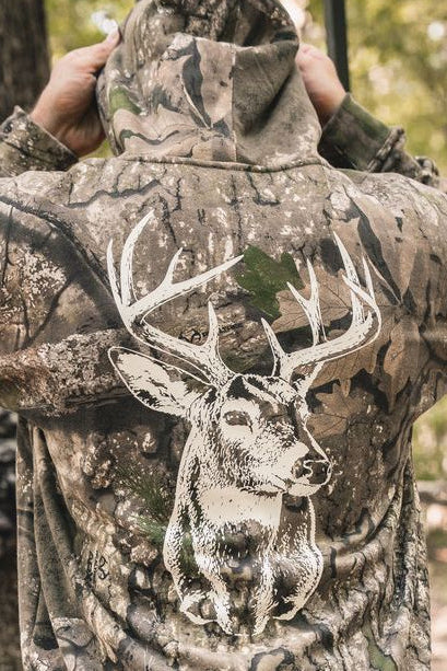 A person wearing The Rut Realtree Premium Hoodie, featuring a Realtree camo pattern and a large white graphic of a deer on the back.