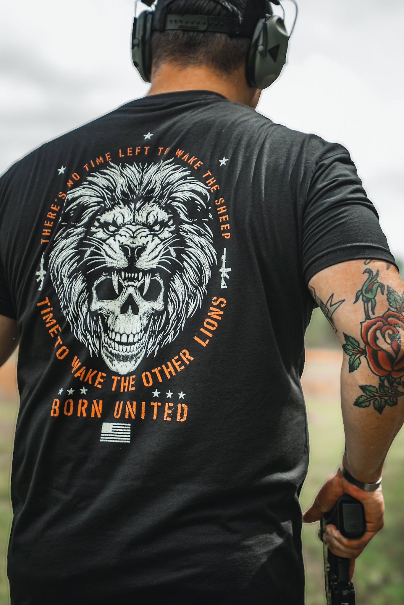Wearing a Waking Lions black T-shirt by Born United Private Label, adorned with a lion and skull graphic and the quote There’s no time left to wake the sheep, time to wake the lions, a man stands outdoors. With headphones, a rose tattoo, and an object in hand, hes ready for whats next.