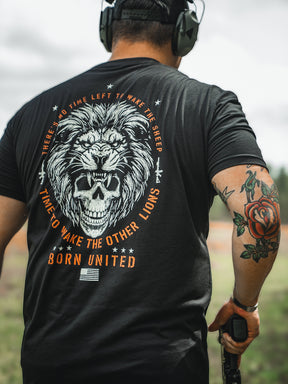 Wearing a Waking Lions black T-shirt by Born United Private Label, adorned with a lion and skull graphic and the quote There’s no time left to wake the sheep, time to wake the lions, a man stands outdoors. With headphones, a rose tattoo, and an object in hand, hes ready for whats next.
