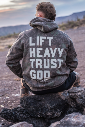 A person sits on rocks in a rugged, mountainous landscape, wearing the Tango Charlie Apparel Lift Heavy. Trust God. - Premium Darkwood Camo Hoodie, which features bold lettering on the back inspired by Proverbs 3:5-6—a powerful scene embodying faith-driven fitness.