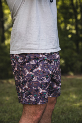 A person outdoors wears a white T-shirt and Mucker Bucket Men's Performance Athletic Shorts | 5.5"- Duck Wild, featuring a playful bird pattern. Blurred greenery in the background focuses attention on their torso and upper legs.