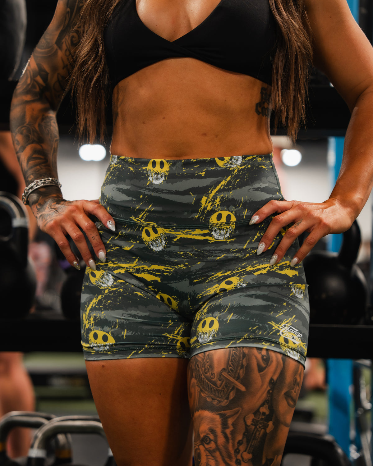 A woman stands in a gym wearing Born United Private Label’s Womens High-Waisted Seamless Front Compression Training Shorts | Unleash the Savage, featuring skull designs. She rests her hands on her hips, highlighting tattoos as gym equipment appears behind her.