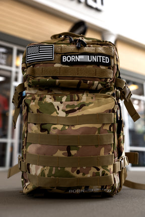 The OCP Tac-Pac - 45L by Born United Private Label, featuring BORN UNITED and American flag patches, stands upright outdoors against a building.