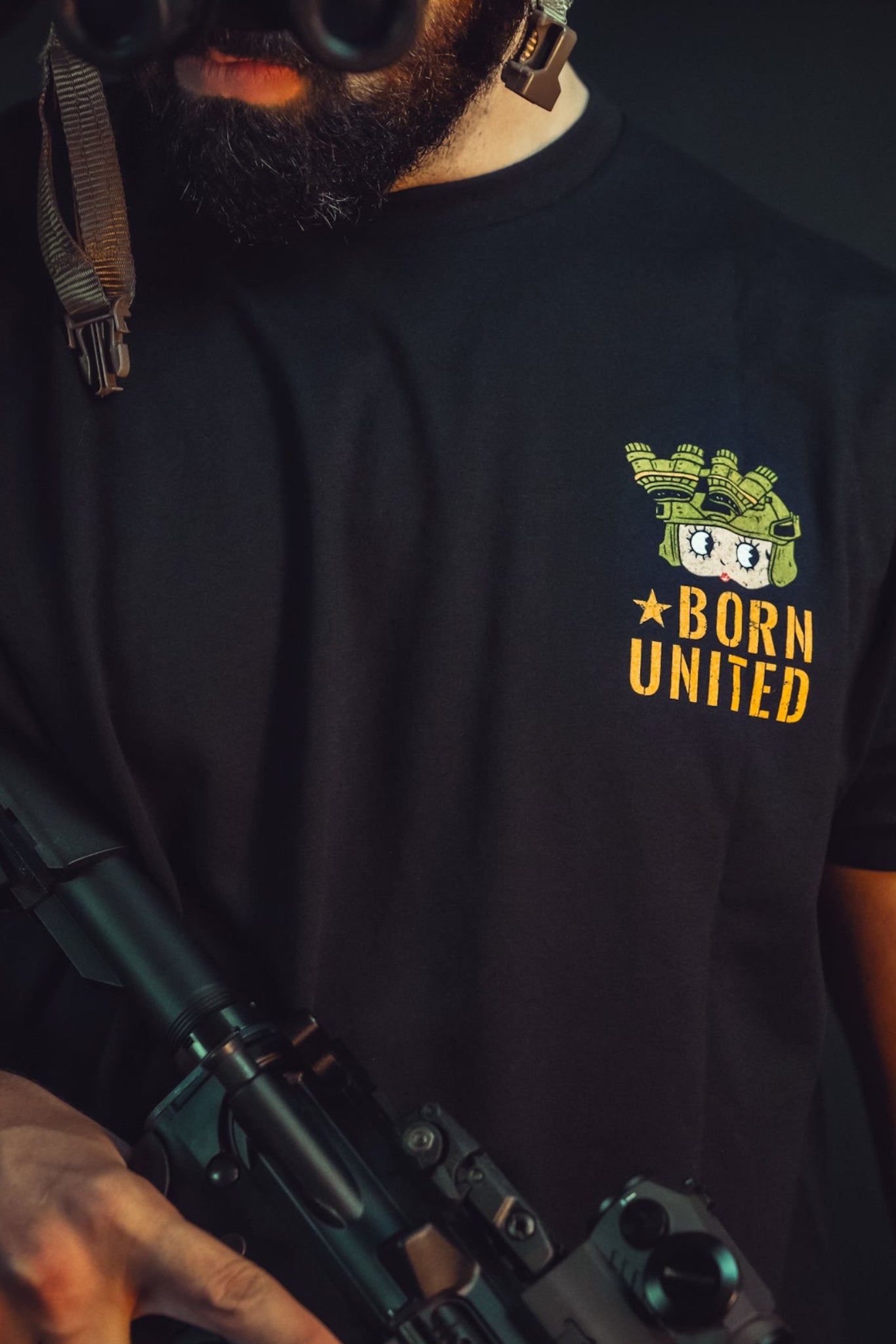 A bearded man in a black Betty Boop Tee by Born United Private Label, symbolizing freedom, grips a rifle. He peers through night vision goggles, tattoos visible on his arm. Defenders like him stand vigilant in the dark background.