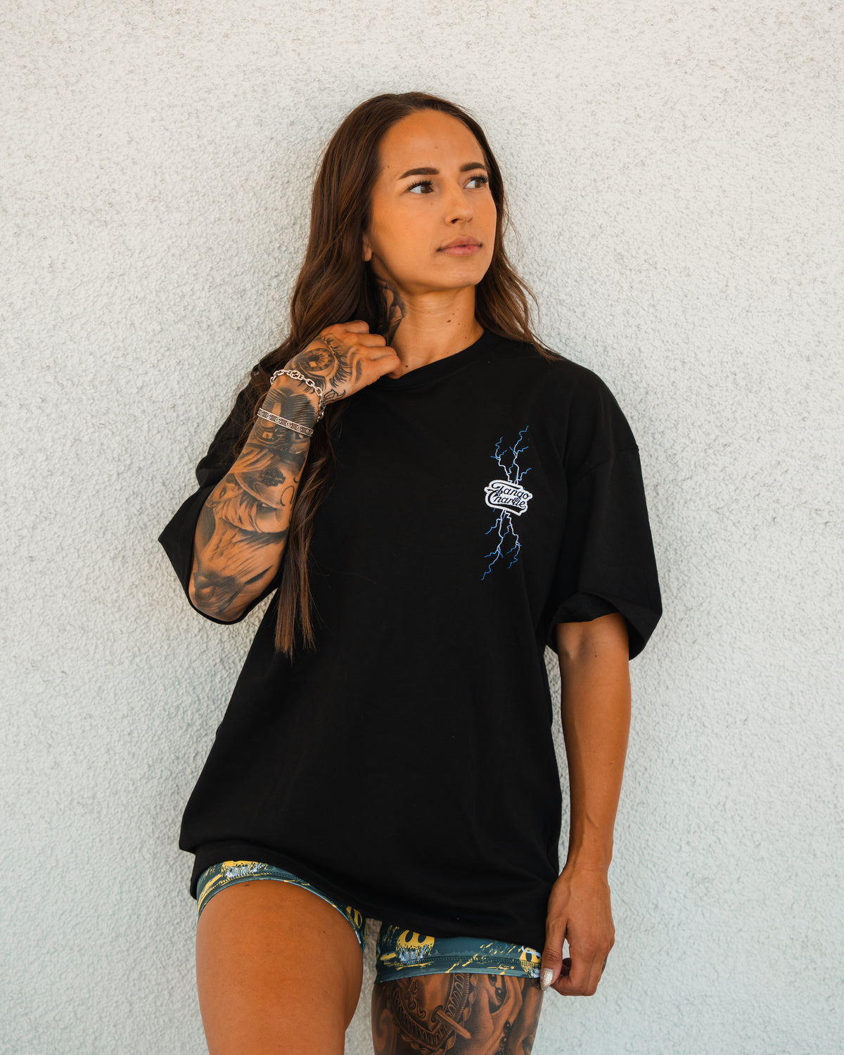 A woman with long brown hair and tattooed arms stands against a light wall, wearing the Tango Charlie Apparel Locked In Premium Oversized Tee in black with a small graphic, paired with patterned shorts, looking to the side.