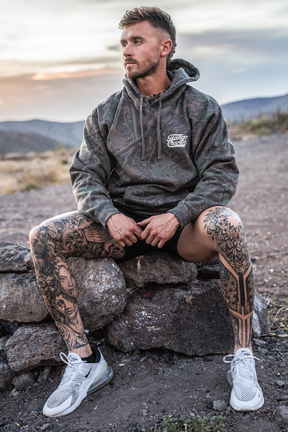 A man with tattooed legs sits on rocks outdoors at sunset, wearing Tango Charlie Apparel’s Lift Heavy. Trust God. - Premium Darkwood Camo Hoodie, black shorts, and white sneakers—a moment capturing the spirit of Christian fitness as he reflects on Proverbs 3:5-6.