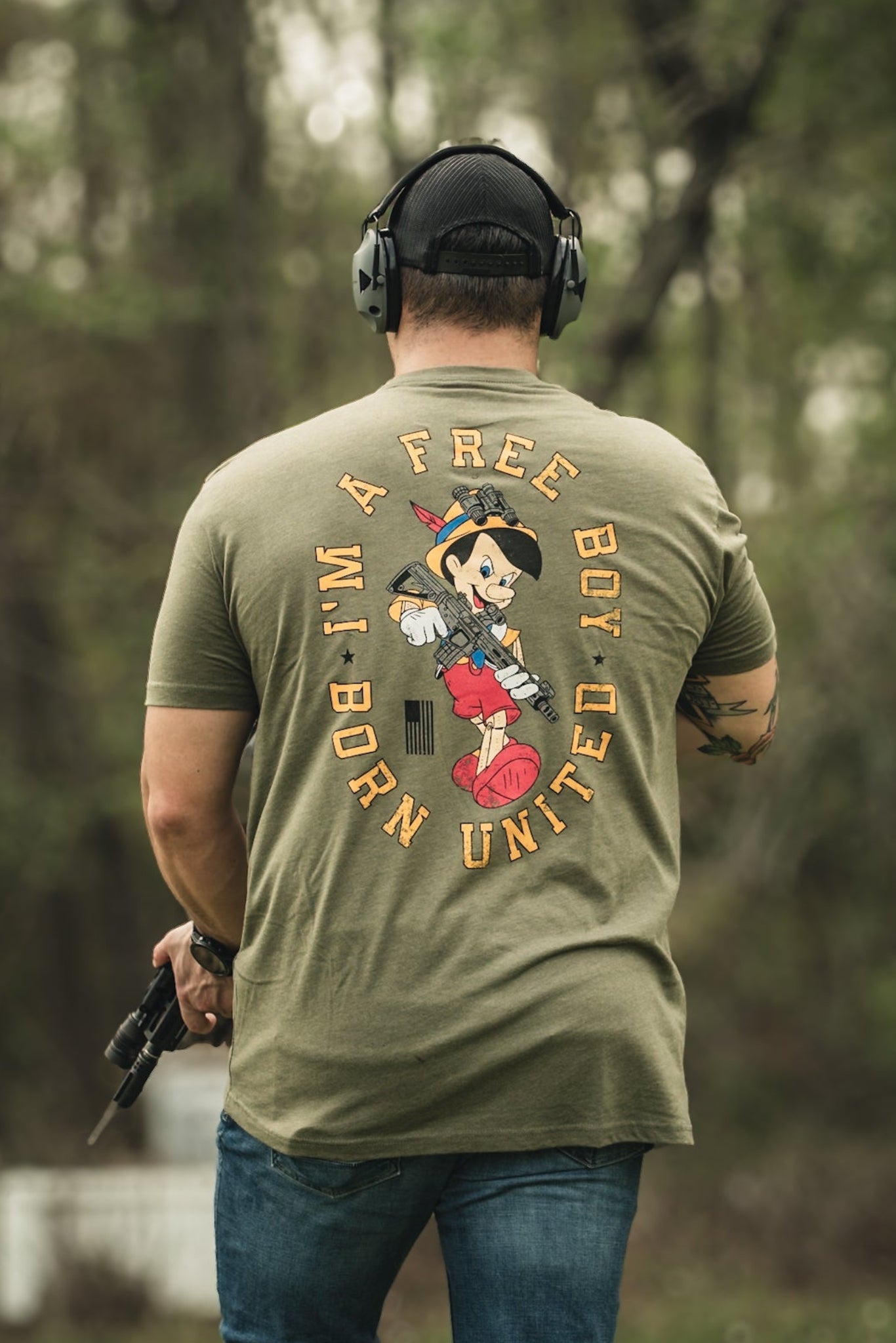 Outdoors, a person wearing headphones and holding a firearm dons the Free Boy Tee by Born United Private Label, featuring an illustrated character with A FREE BOY UNITE text. Lush trees and foliage fill the background.