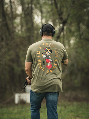 Outdoors, a person wearing headphones and holding a firearm dons the Free Boy Tee by Born United Private Label, featuring an illustrated character with A FREE BOY UNITE text. Lush trees and foliage fill the background.