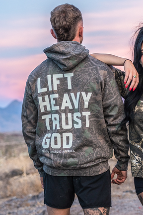 A person is wearing the Tango Charlie Apparel Lift Heavy. Trust God. - Premium Darkwood Camo Hoodie, featuring Christian fitness inspiration from Proverbs 3:5-6, while standing outdoors with another individual partially visible.