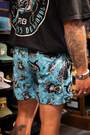 A person with tattooed arms wears a black t-shirt and Relentless Betrayal Premium Mesh Shorts | 5.5"- Davy Jones, which feature bold tattoo-inspired designs, standing in a hat store with hats displayed on shelves in the background.