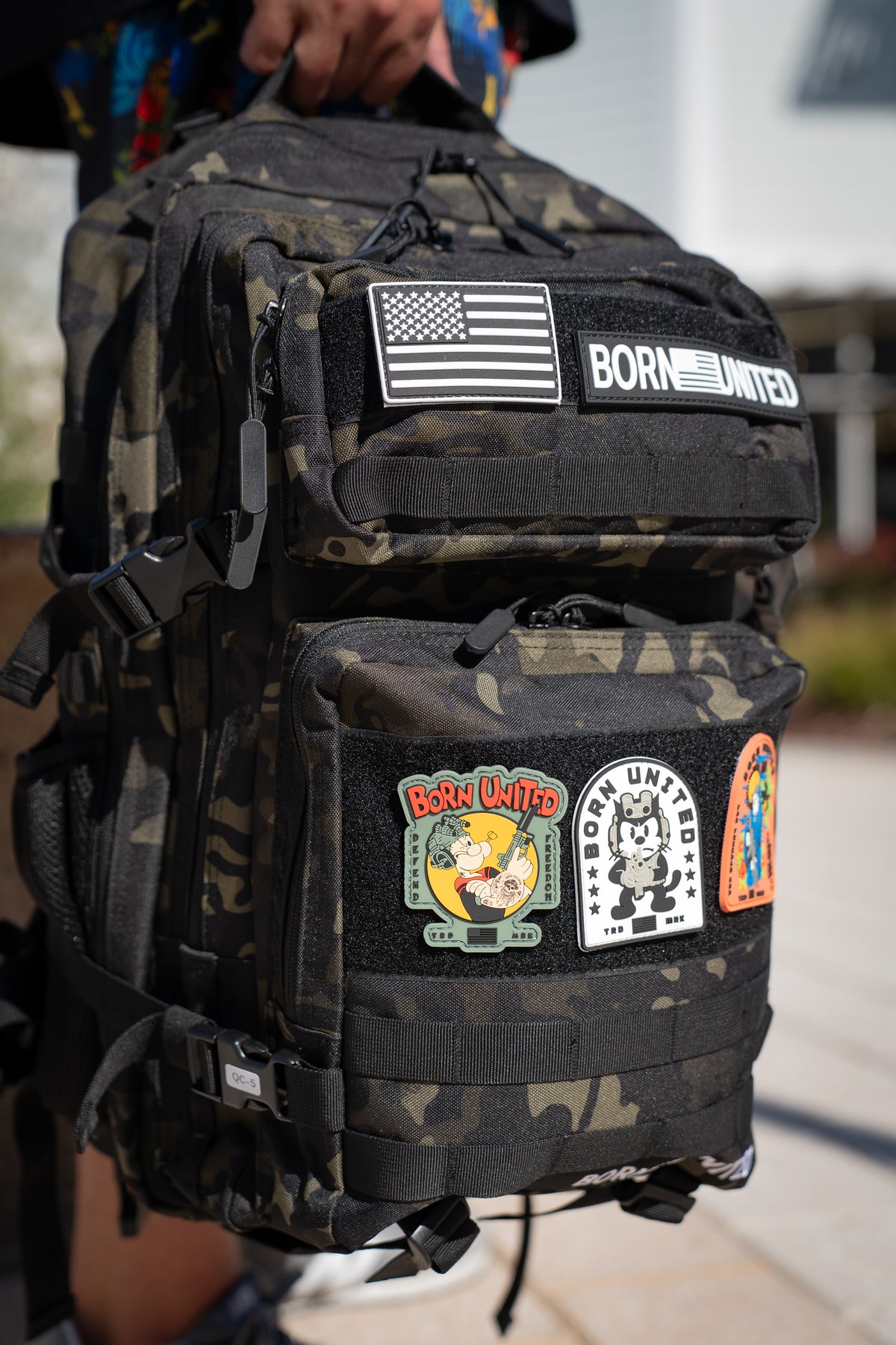 A person holds the Born United Private Label Black Camo Premium Tac-Pac - 45L outdoors, featuring colorful patches like a U.S. flag, BORN UNITED, and cartoon designs.