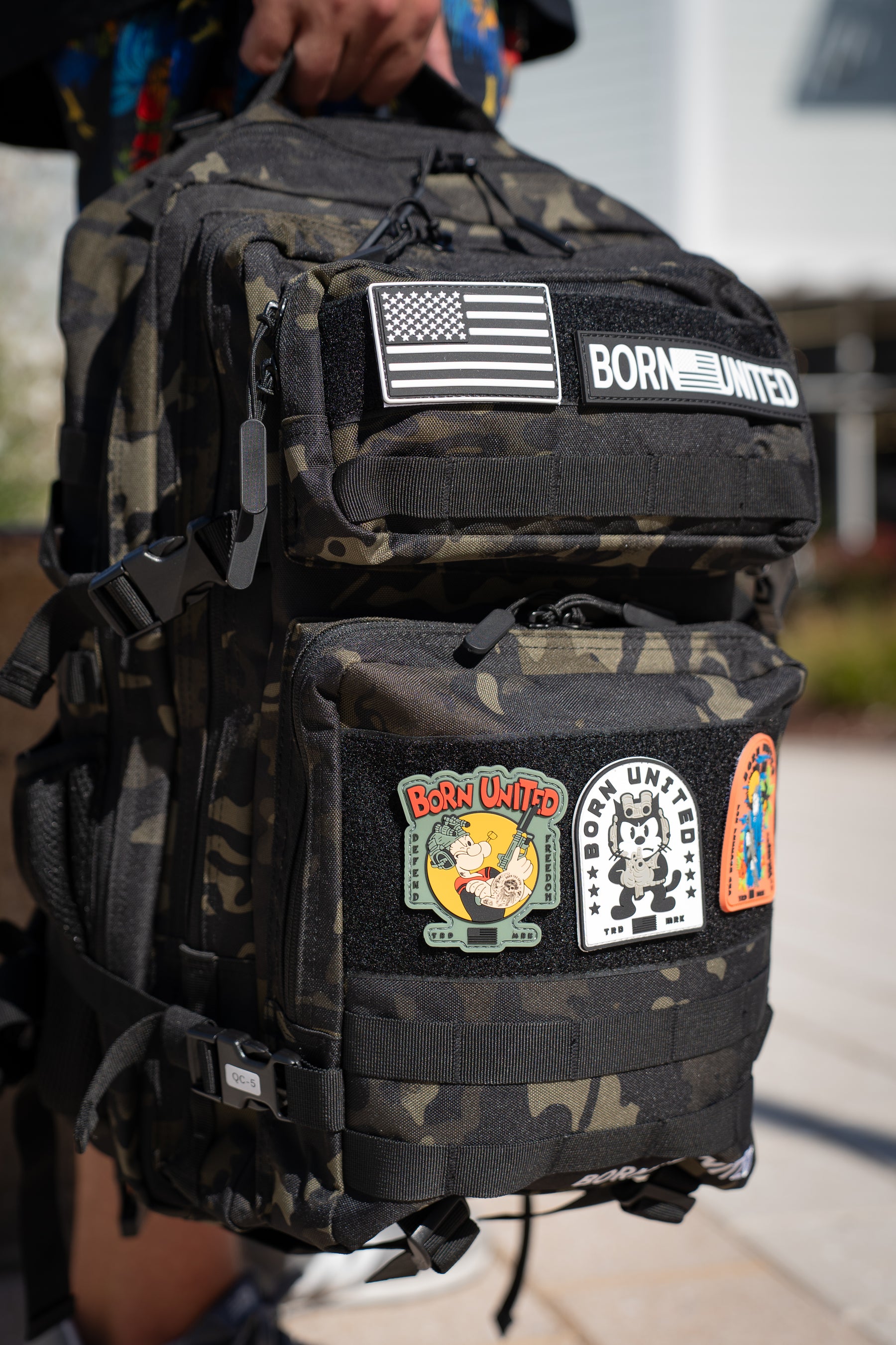 A person holds the Born United Private Label Black Camo Premium Tac-Pac - 45L outdoors, featuring colorful patches like a U.S. flag, BORN UNITED, and cartoon designs.