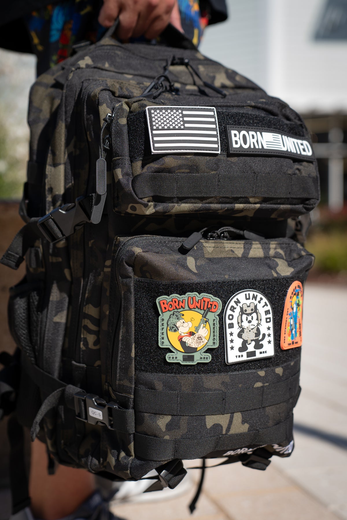 Outdoors in daylight, a person holds the Black Camo Premium Tac-Pac - 45L by Born United Private Label, featuring several colorful patches such as an American flag, BORN UNITED, and cartoon characters.