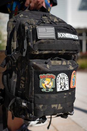 Outdoors in daylight, a person holds the Black Camo Premium Tac-Pac - 45L by Born United Private Label, featuring several colorful patches such as an American flag, BORN UNITED, and cartoon characters.