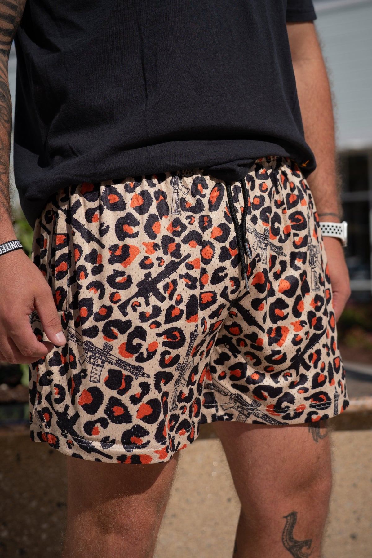 A tattooed person stands outdoors in a black T-shirt and Tango Charlie Apparel's Men's Mesh Lifestyle Shorts | 5.5"- Loaded Leopard, featuring a bold leopard print, black wristband, and white watch—perfect for any stylish occasion.