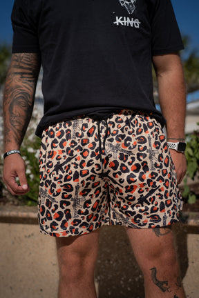 A person stands outdoors in a black KING t-shirt and Tango Charlie Apparel Men's Mesh Lifestyle Shorts | 5.5"- Loaded Leopard. Tattooed arms, a wristband, and checkered watch pop against blurred greenery for a bold lifestyle shorts look.