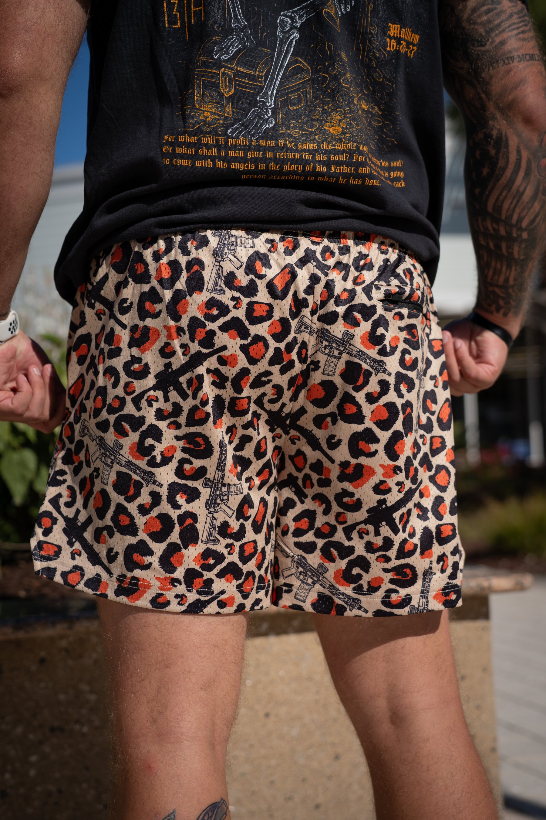 Outdoors, a person wears Tango Charlie Apparel's Men's Mesh Lifestyle Shorts | 5.5"- Loaded Leopard—leopard print with Eiffel Tower accents—and a black tee with orange/yellow text, showing arm and leg tattoos amid buildings and greenery.