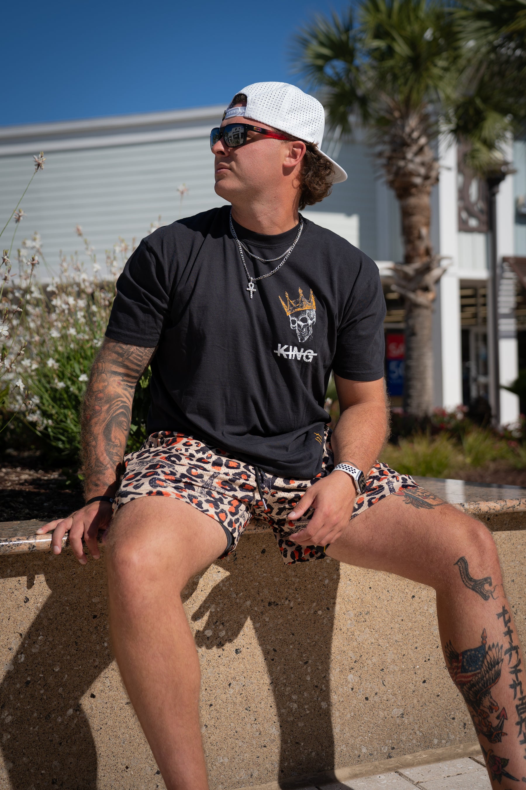 A man wearing Tango Charlie Apparel’s Men's Mesh Lifestyle Shorts | 5.5"–Loaded Leopard sits on a low wall outdoors. He sports a white backward cap, black graphic tee, sunglasses, tattoos, necklaces, and a watch.
