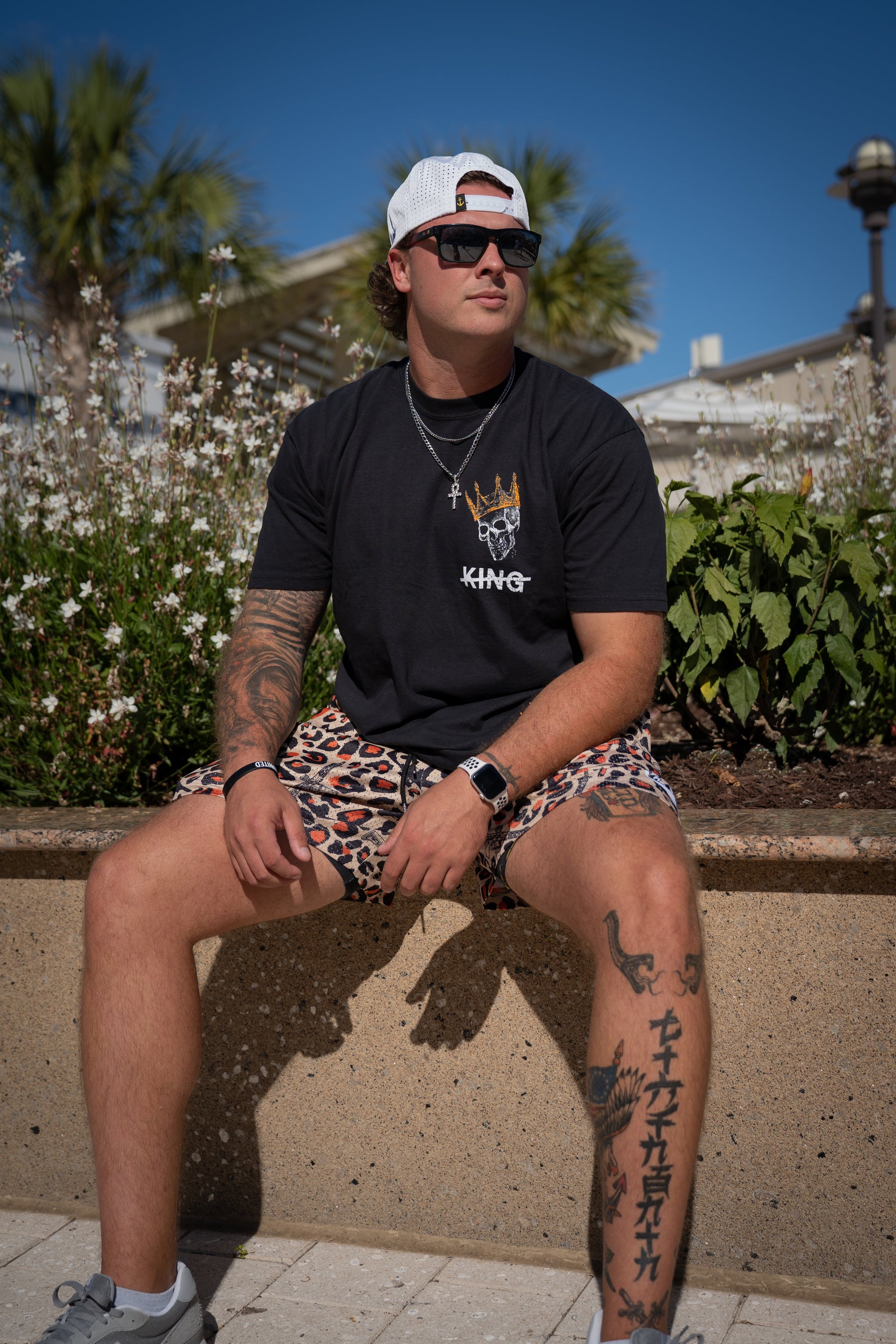 A man in a white cap, sunglasses, black t-shirt, and Tango Charlie Apparel Men's Mesh Lifestyle Shorts | 5.5" - Loaded Leopard sits on a low wall outdoors with flowers and palm trees under a blue sky.