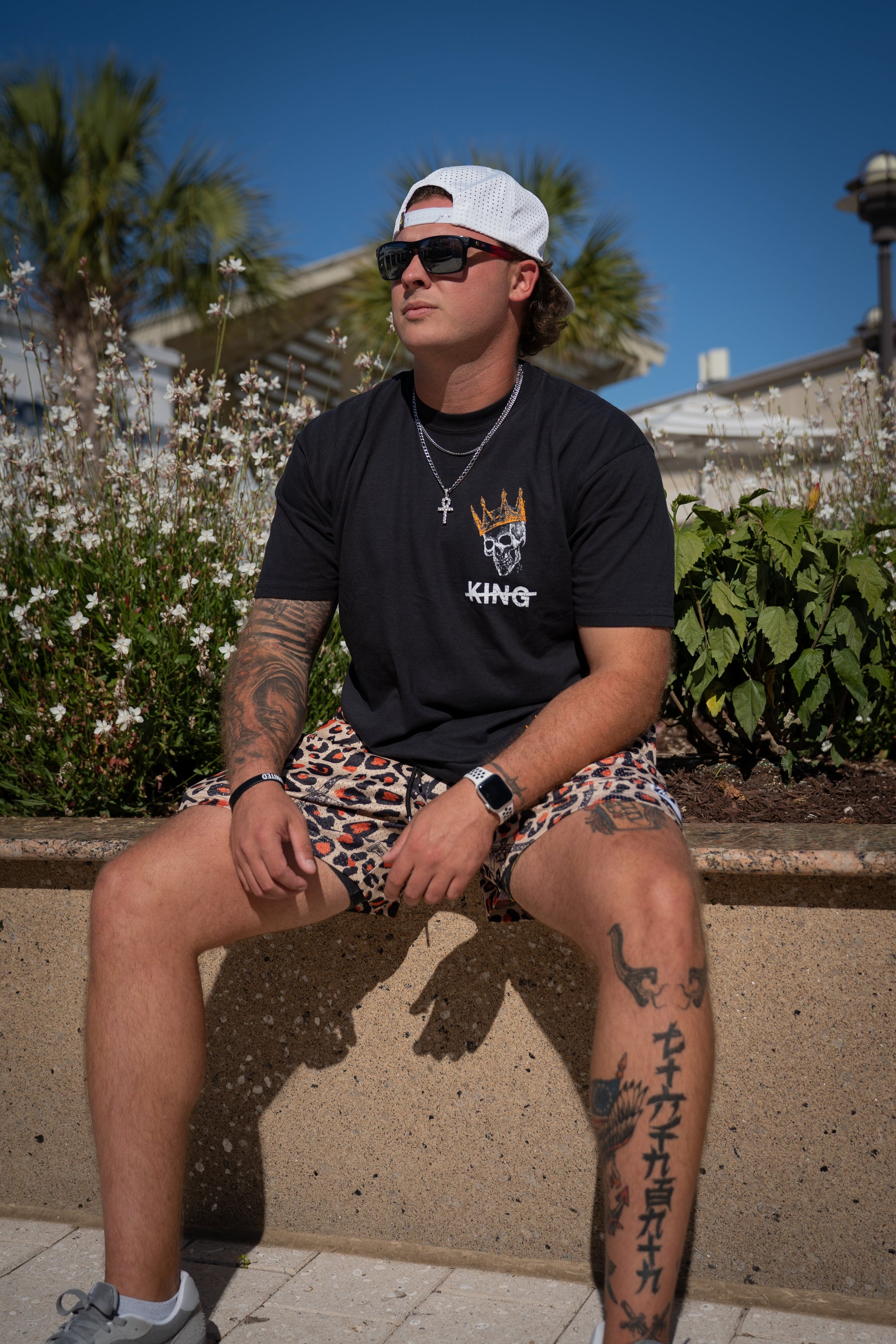 A man sporting sunglasses, a white cap, a black graphic tee, and Tango Charlie Apparel’s Men's Mesh Lifestyle Shorts | 5.5"- Loaded Leopard sits on a low wall outdoors, showing arm and leg tattoos amid greenery and flowers.