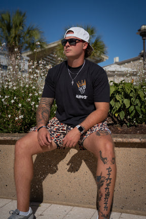 A man sporting sunglasses, a white cap, a black graphic tee, and Tango Charlie Apparel’s Men's Mesh Lifestyle Shorts | 5.5"- Loaded Leopard sits on a low wall outdoors, showing arm and leg tattoos amid greenery and flowers.
