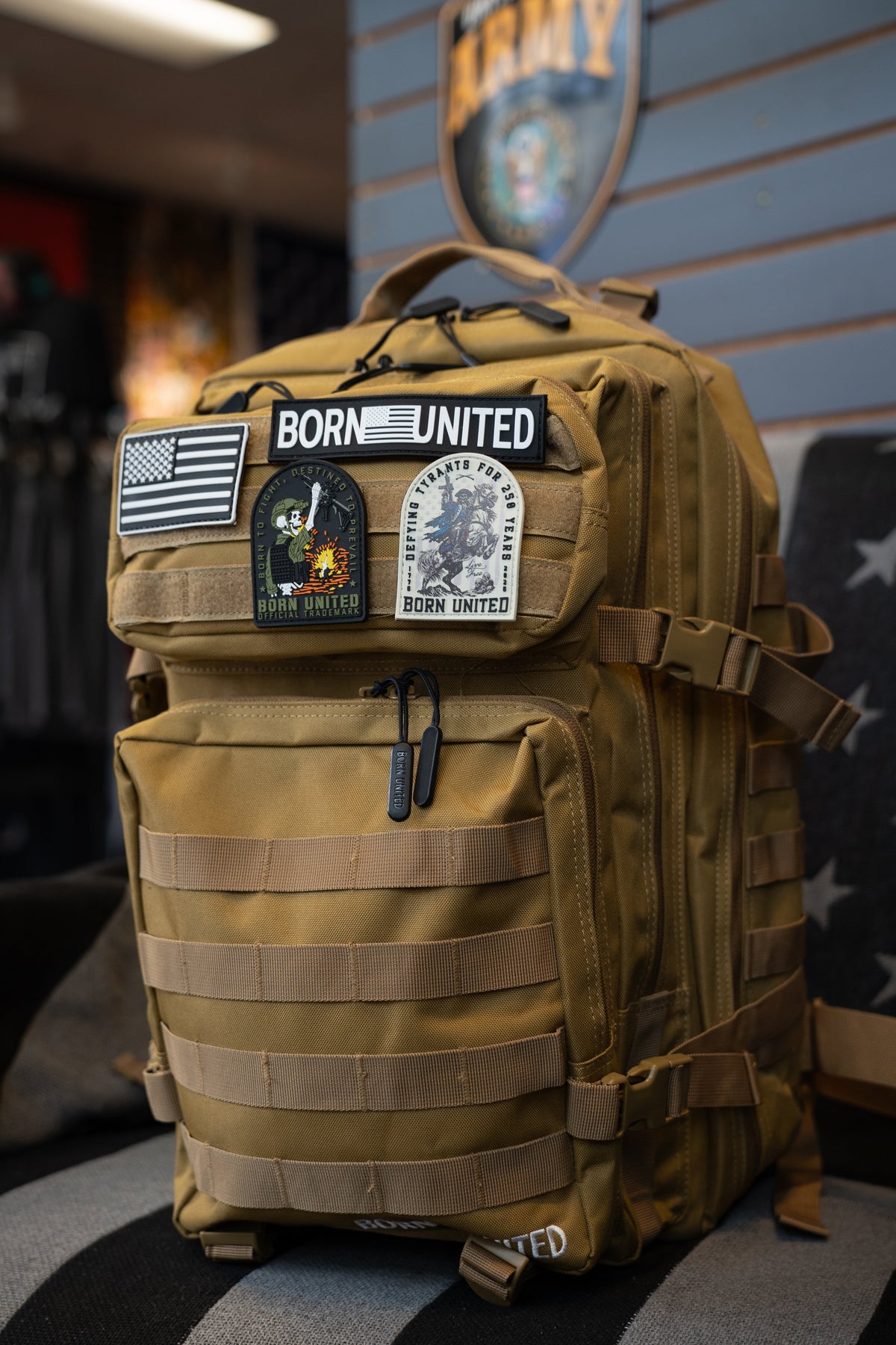 The Coyote Brown Tac-Pac - 45L by Born United Private Label, featuring multiple patches such as BORN UNITED and an American flag, is displayed on a couch in a military-themed store.