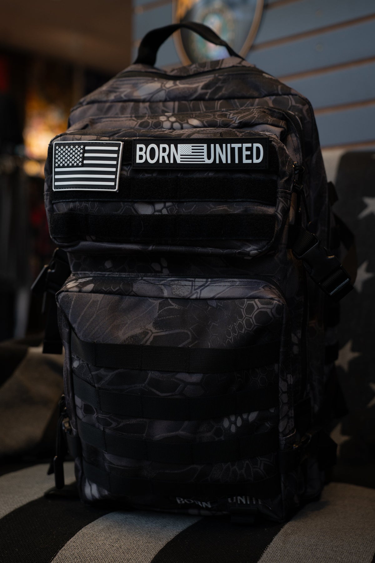 A black tactical backpack with a dark gray camouflage pattern, featuring a front pocket with a black and white American flag patch and a 'BORN UNITED' logo, resting on a striped black and gray surface.