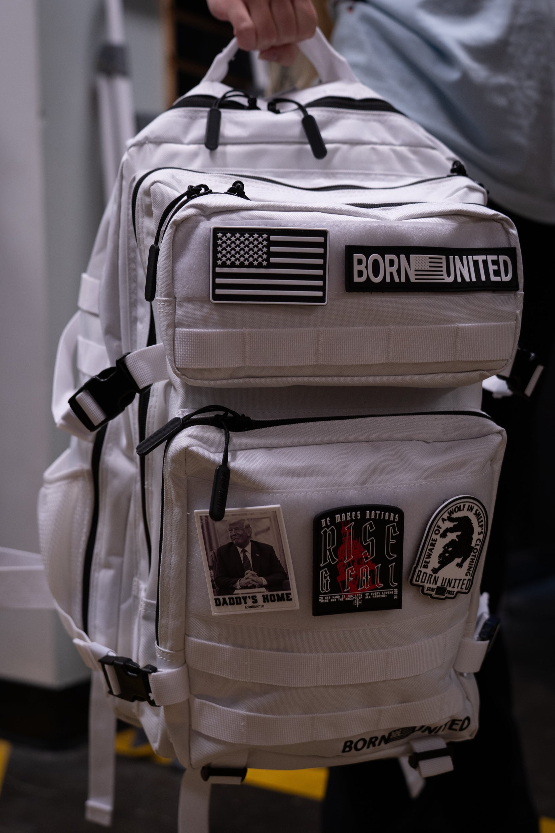 A white tactical-style backpack with black straps and multiple patches, including a prominent 'BORN UNITED' patch, being held by a person in a gray shirt.