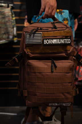 A person holds the Chocolate Tac-Pac - 25L by Born United Private Label, featuring black and white American flag and BORN UNITED patches, while standing in a store. Designed for organized storage, this tactical backpack seamlessly blends functionality with style.