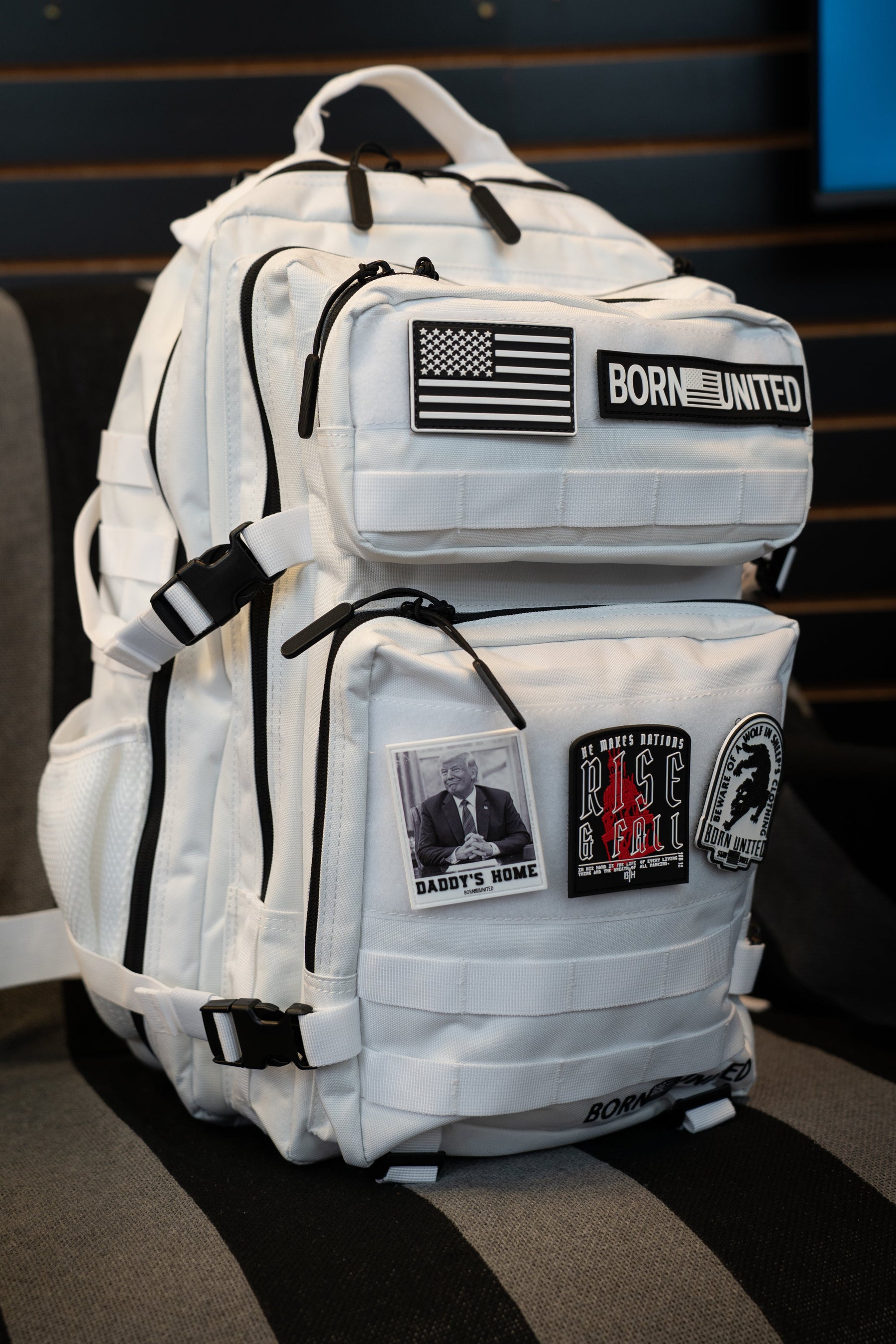A white tactical backpack with black straps and multiple patches, including a 'Born United' patch, set on a dark striped chair.