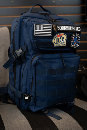The Navy Blue Tac-Pac - 45L by Born United Private Label, adorned with multiple patches such as a U.S. flag, "BORN UNITED," and beer-themed designs, sits on a chair. Its rich Navy Blue color enhances the rugged style and distinctive appearance of this tactical backpack.