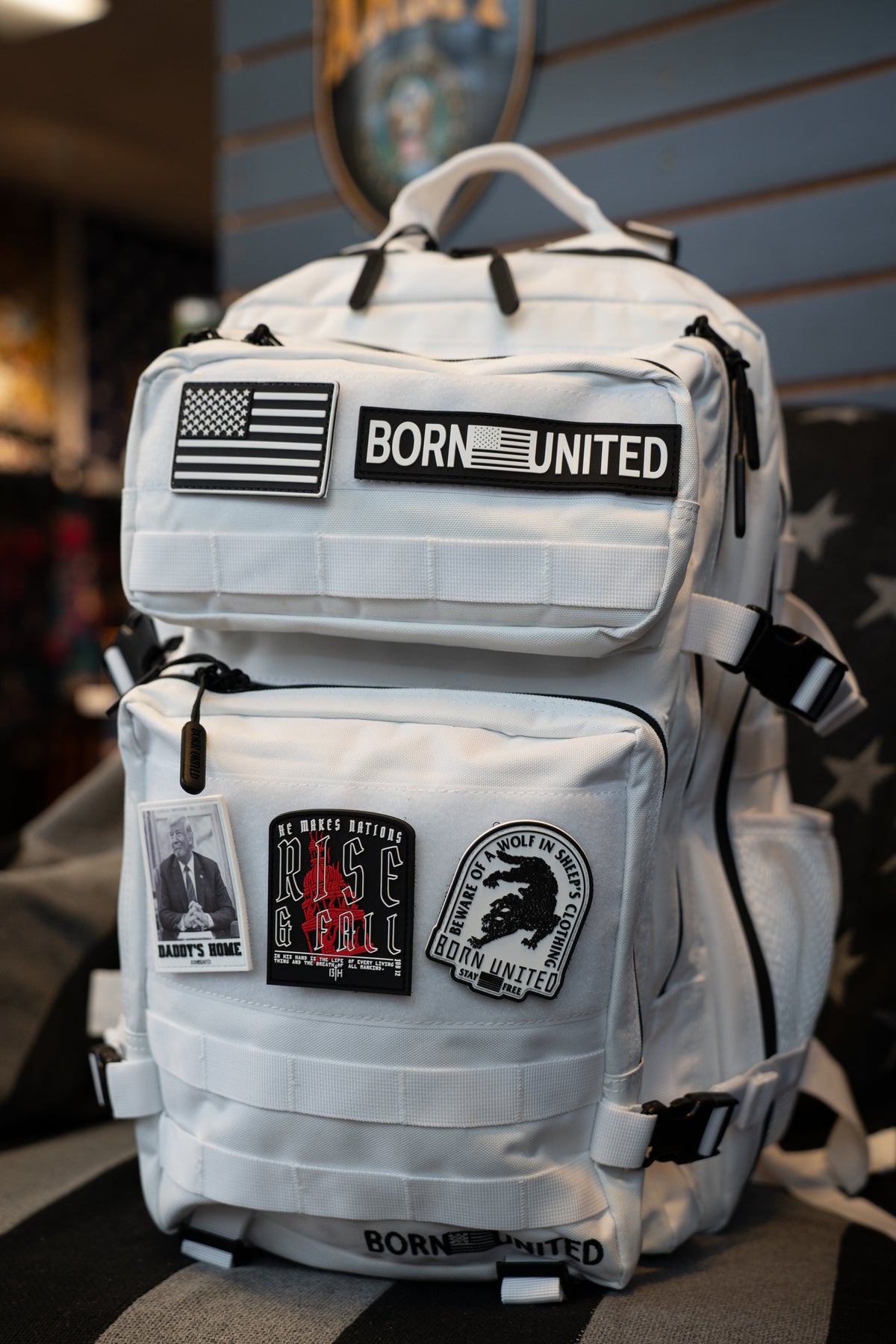 A white tactical backpack with multiple patches, including a 'BORN UNITED' patch and an American flag patch, resting on a striped black and gray surface.
