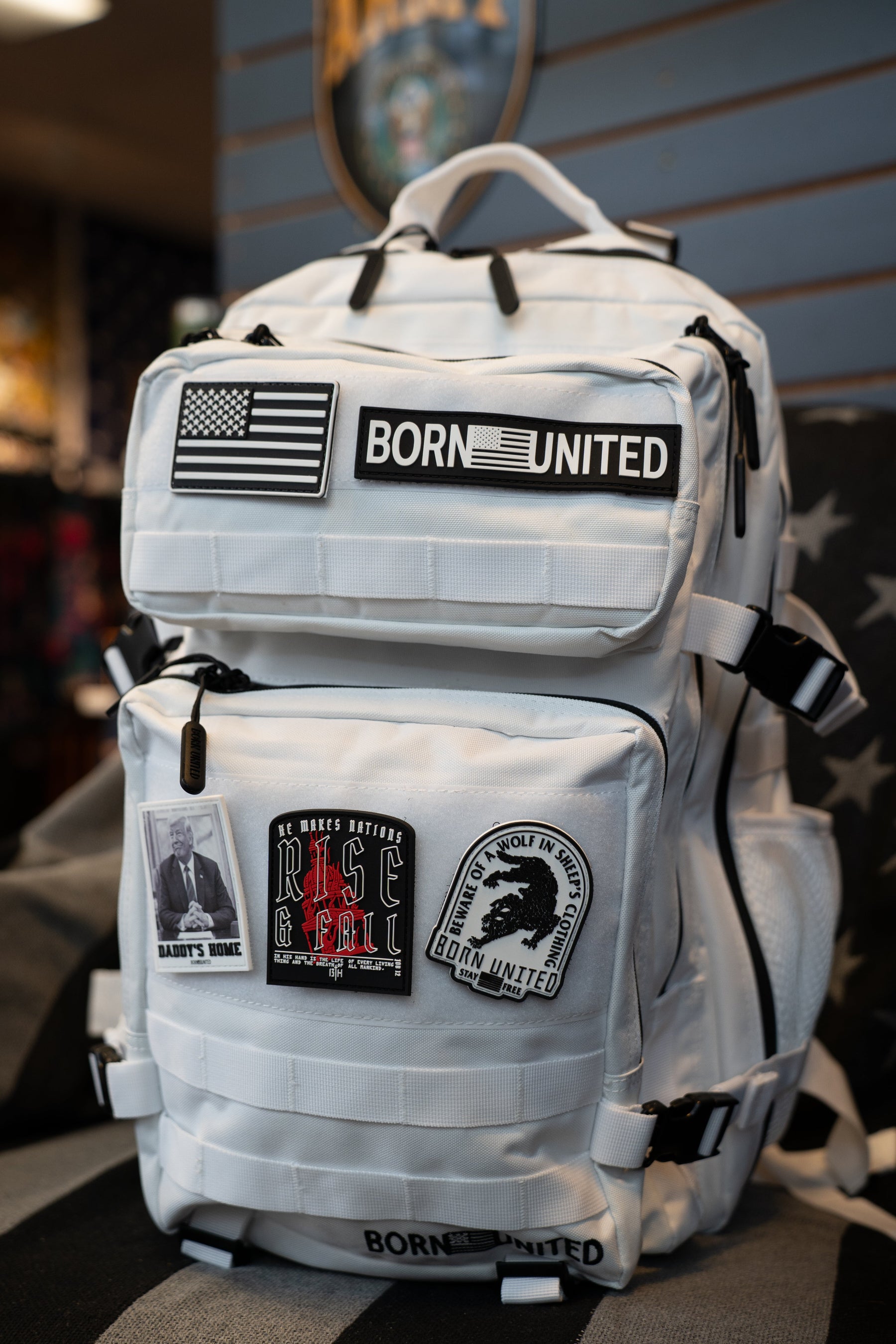 A white tactical backpack with multiple patches, including a 'BORN UNITED' patch and an American flag patch, resting on a striped black and gray surface.