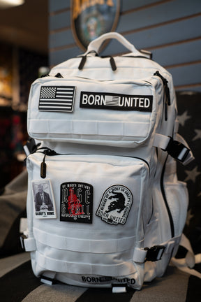 A white tactical backpack with multiple patches, including a 'BORN UNITED' patch and an American flag patch, resting on a striped black and gray surface.