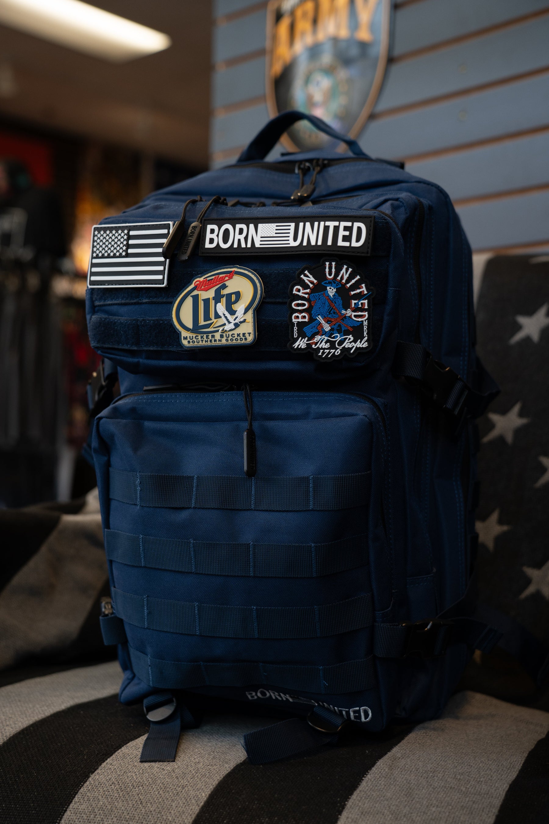 The Navy Blue Tac-Pac - 45L by Born United Private Label showcases multiple patches—including BORN UNITED, a US flag, and LIFE—and is displayed indoors on a star-adorned striped surface.