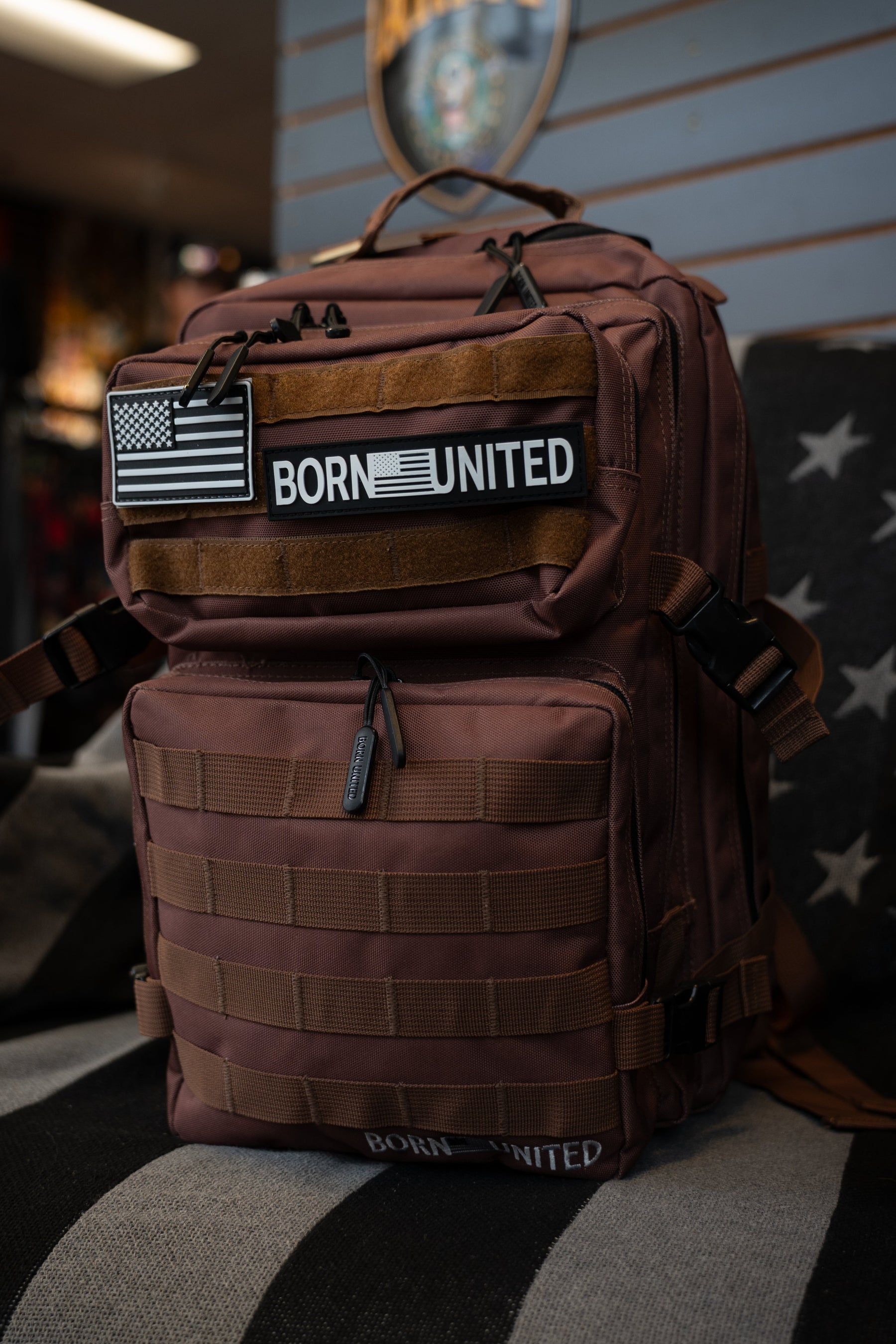 The Chocolate Tac-Pac - 25L by Born United Private Label, adorned with American flag and BORN UNITED patches, features organized storage and is displayed indoors on a star-patterned chair.