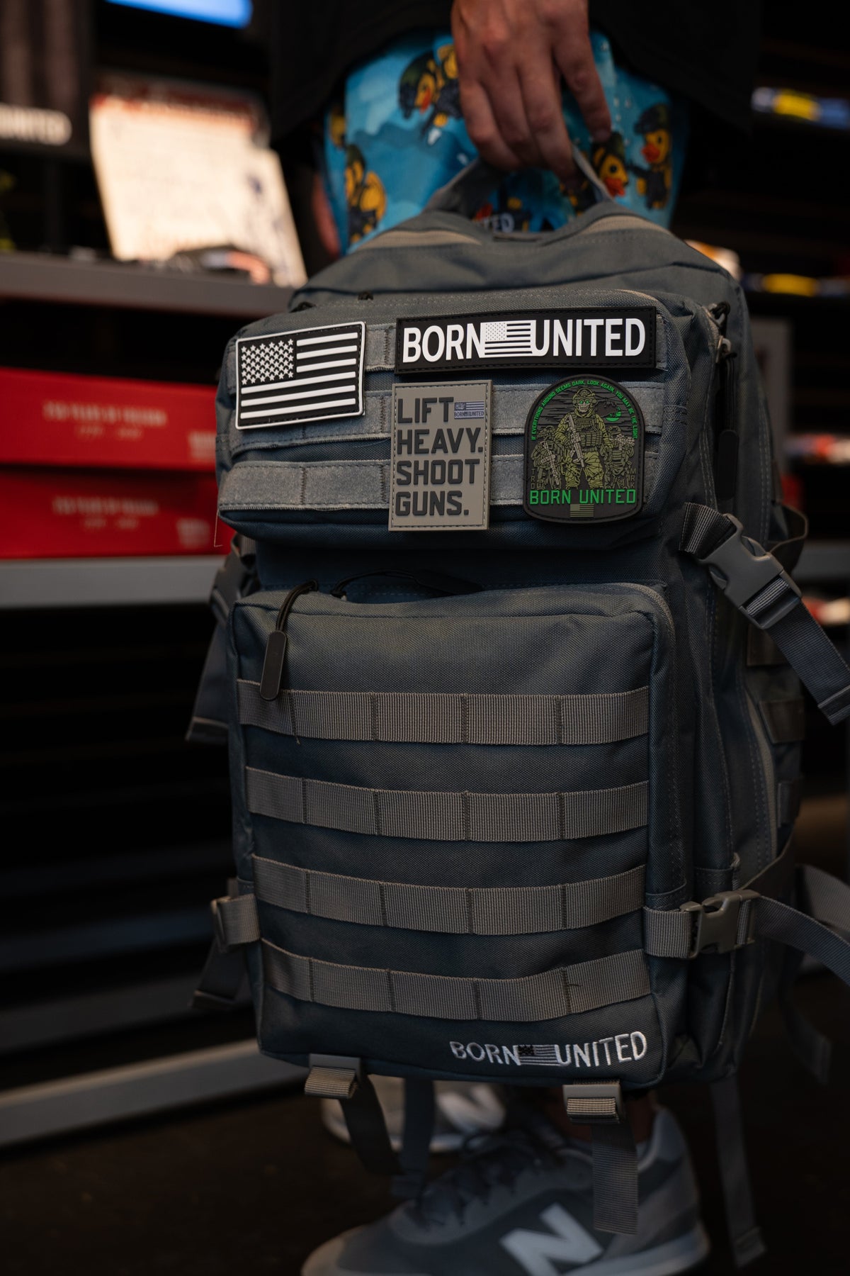 A person holds a Born United Private Label Pilot Gray Tac-Pac - 45L backpack, adorned with various patches such as BORN UNITED, an American flag, LIFT HEAVY. SHOOT GUNS., and a camouflage patch.