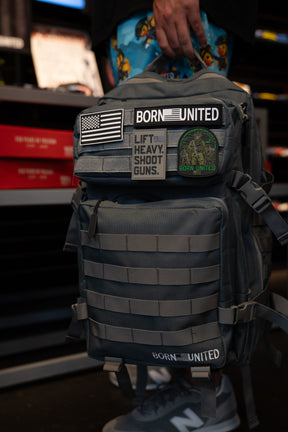 A person holds a Born United Private Label Pilot Gray Tac-Pac - 45L backpack, adorned with various patches such as BORN UNITED, an American flag, LIFT HEAVY. SHOOT GUNS., and a camouflage patch.