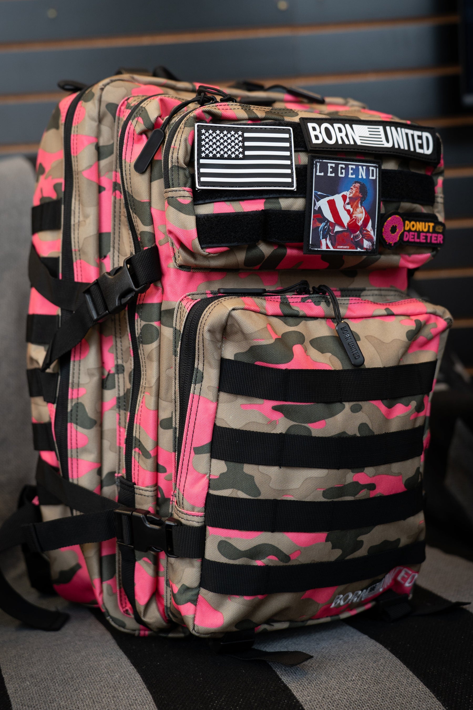 A pink and tan camouflage backpack with black straps and multiple utility pockets, featuring a 'Born United' patch on the front, set against a dark wood slat wall background.