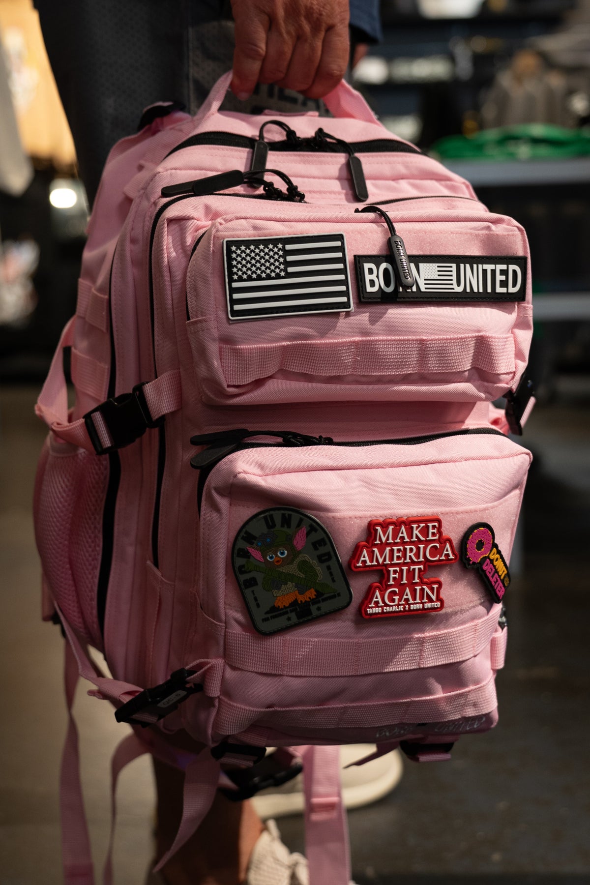 A hand holds the Pink Mist Tac-Pac - 25L by Born United Private Label, a vibrant pink backpack decorated with multiple patches—an American flag, "BORN UNITED," "MAKE AMERICA FIT AGAIN," and a fun donut patch. This 25-liter travel backpack combines standout style and practical function for all your daily adventures.