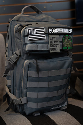 A Born United Private Label Pilot Gray Tac-Pac - 45L tactical backpack with MOLLE webbing features several patches, including an American flag, “Born United,” “Lift Heavy. Shoot Guns.,” and a forest design patch.