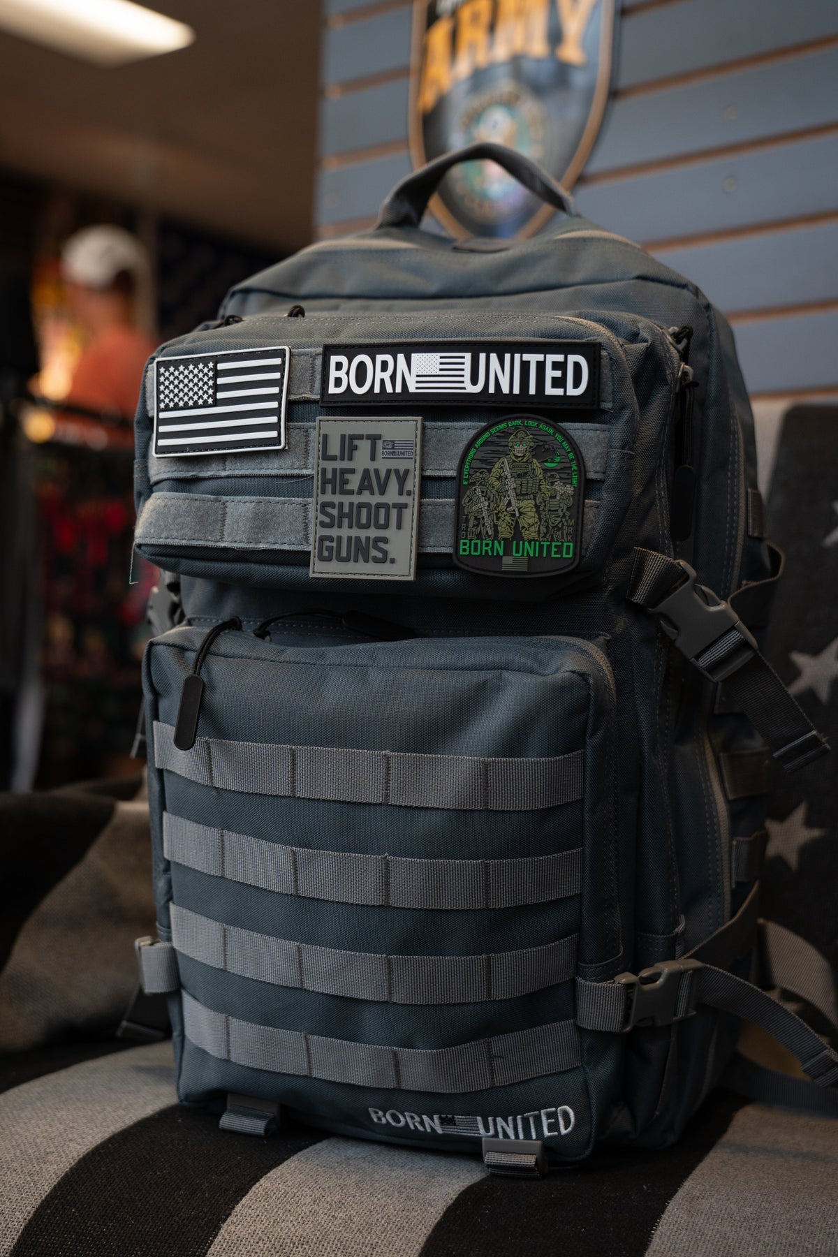 A Born United Private Label Pilot Gray Tac-Pac - 45L, featuring multiple patches such as an American flag, BORN UNITED, and LIFT HEAVY. SHOOT GUNS., sits on a chair indoors.