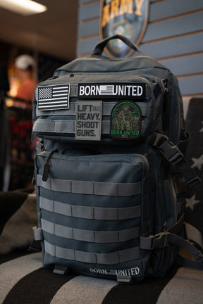 A Born United Private Label Pilot Gray Tac-Pac - 45L, featuring multiple patches such as an American flag, BORN UNITED, and LIFT HEAVY. SHOOT GUNS., sits on a chair indoors.